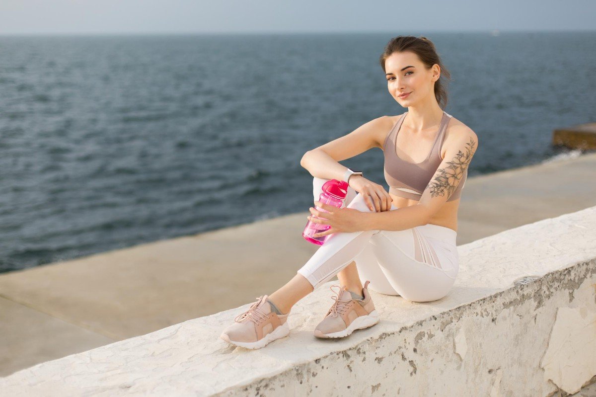 mulher com roupas de academia sentada em mureta na frente do mar