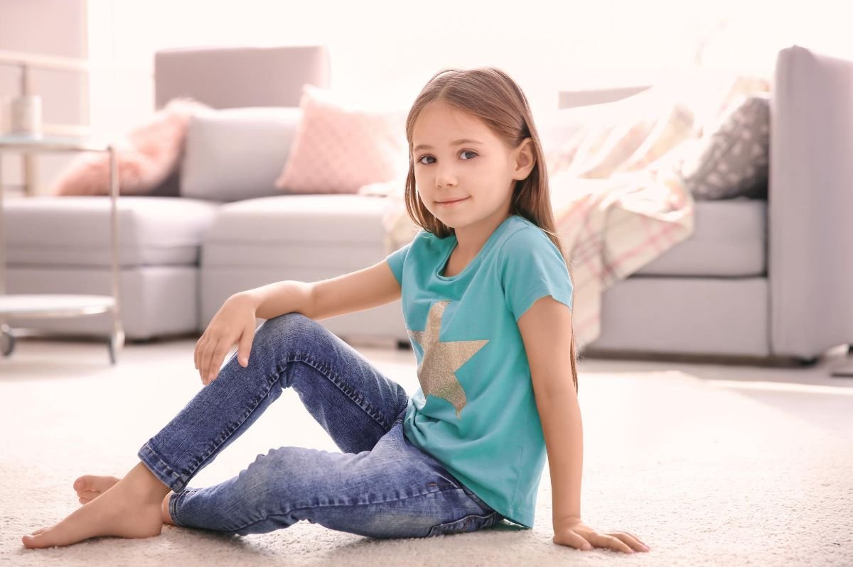 menina sentada no chão de casa usando camisa verde e calcas infantis