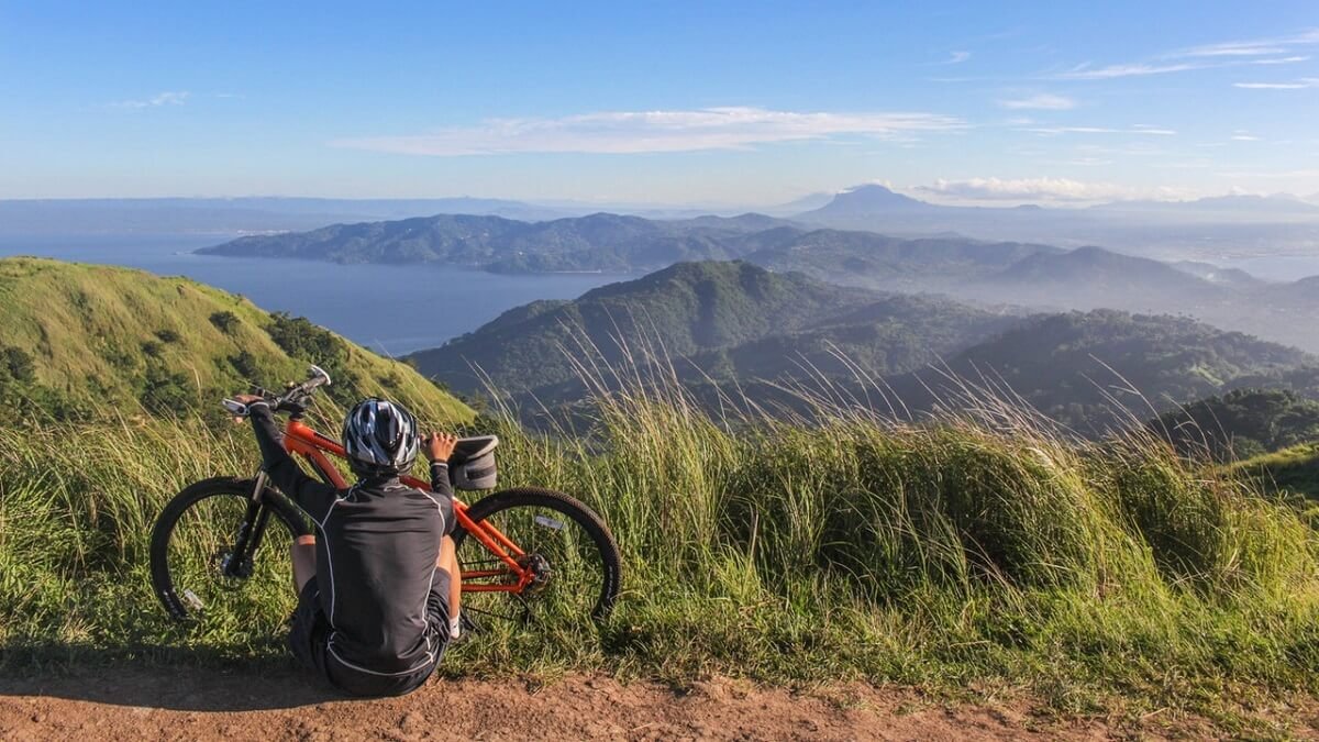 Homem sentado junto com a sua bicicleta em lugar alto com montanhas ao fundo